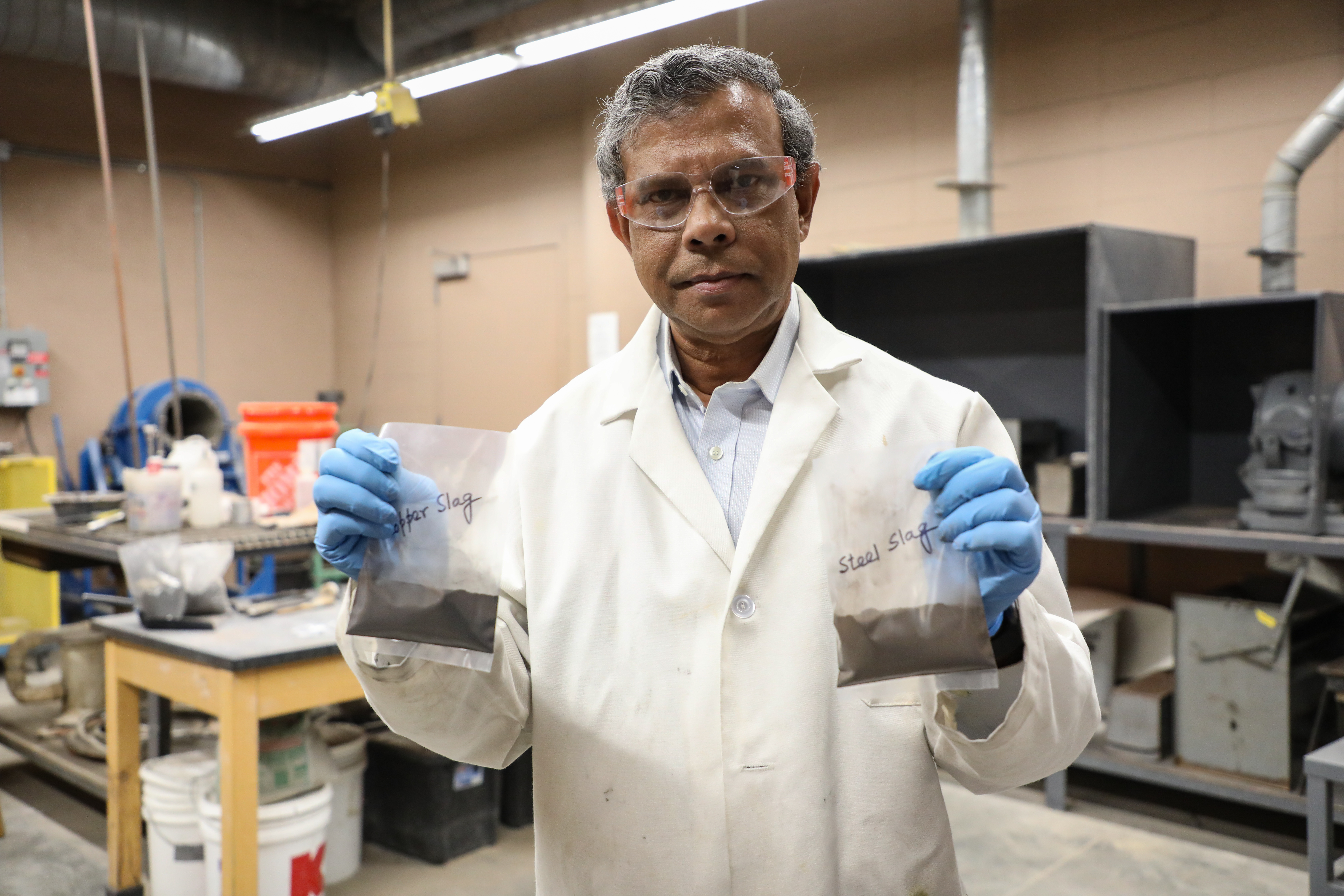!Professor Das holds two see-through baggies, one marked "copper slag" the other marked "steel slag."
