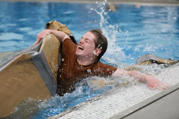 A student paddles in the pool, clinging to a sunken cardboard boat. 