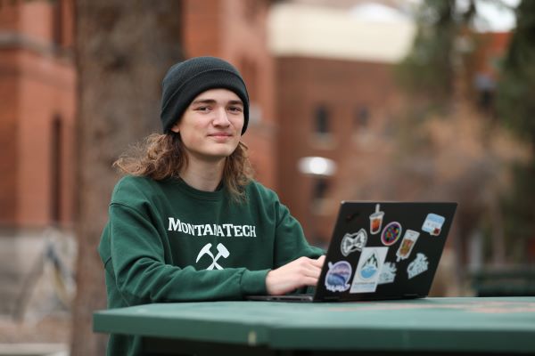 Nathan Rayburn in a green Montana Tech sweatshirt, sitting at a table with a laptop covered in stickers