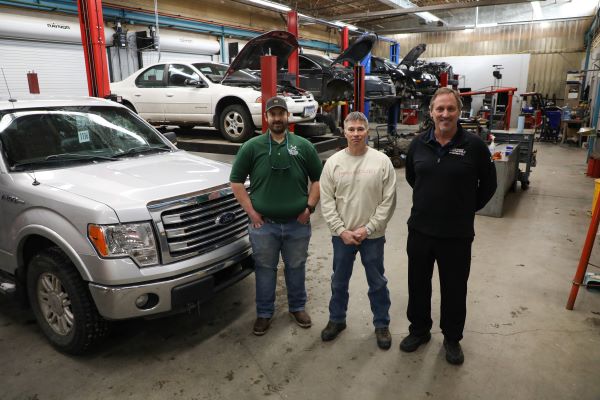 Three men stand in front of vehicles in a workshop