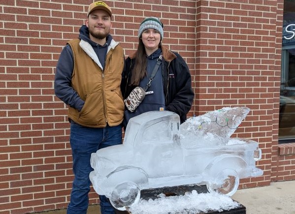 !Ethan and Aubrey Teller stand in front of an ice sculpture of a pickup truck carrying a Christmas tree