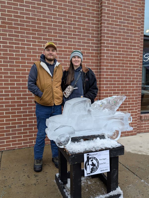 Ethan and Aubrey Teller stand in front of an ice sculpture of a pickup truck carrying a Christmas tree